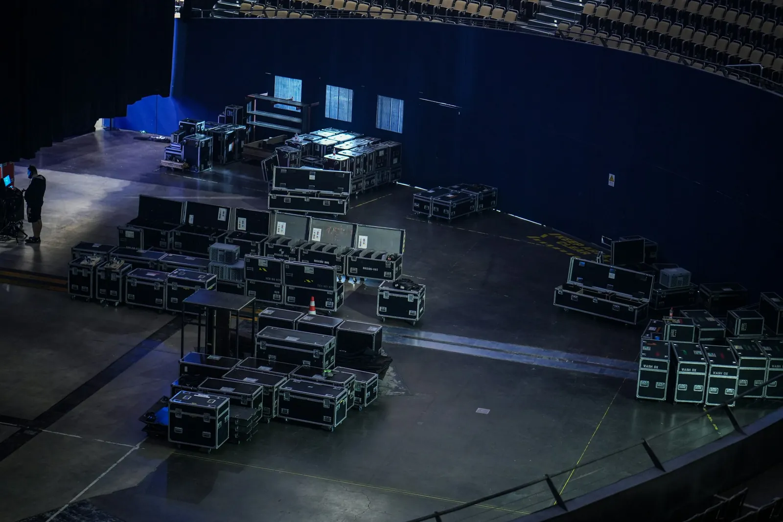Rows of equipment road cases laid out on the floor of a large arena before an event, with a technician standing at the left