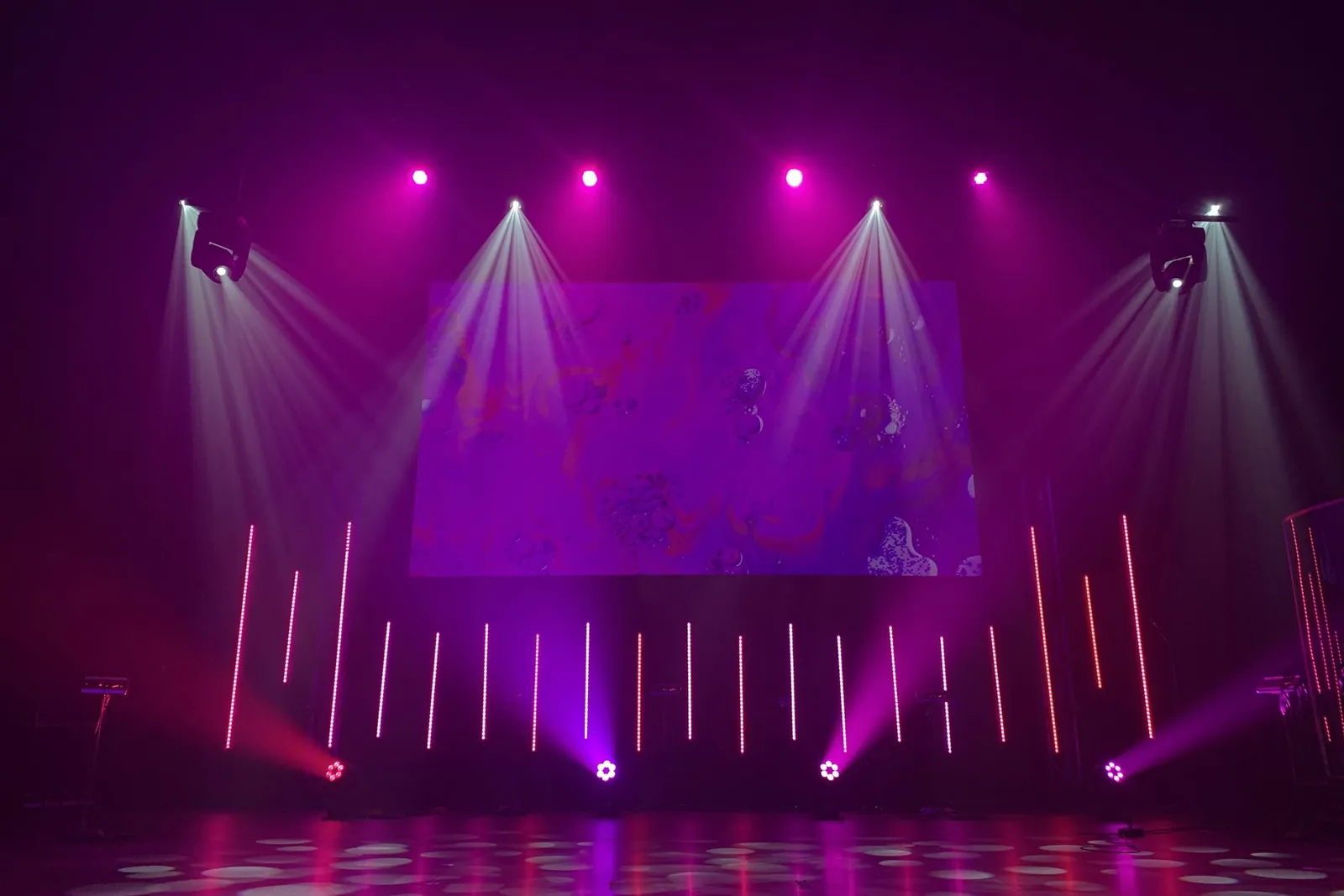 Stage lit in purple and magenta with spotlights, vertical light bars and a projection screen